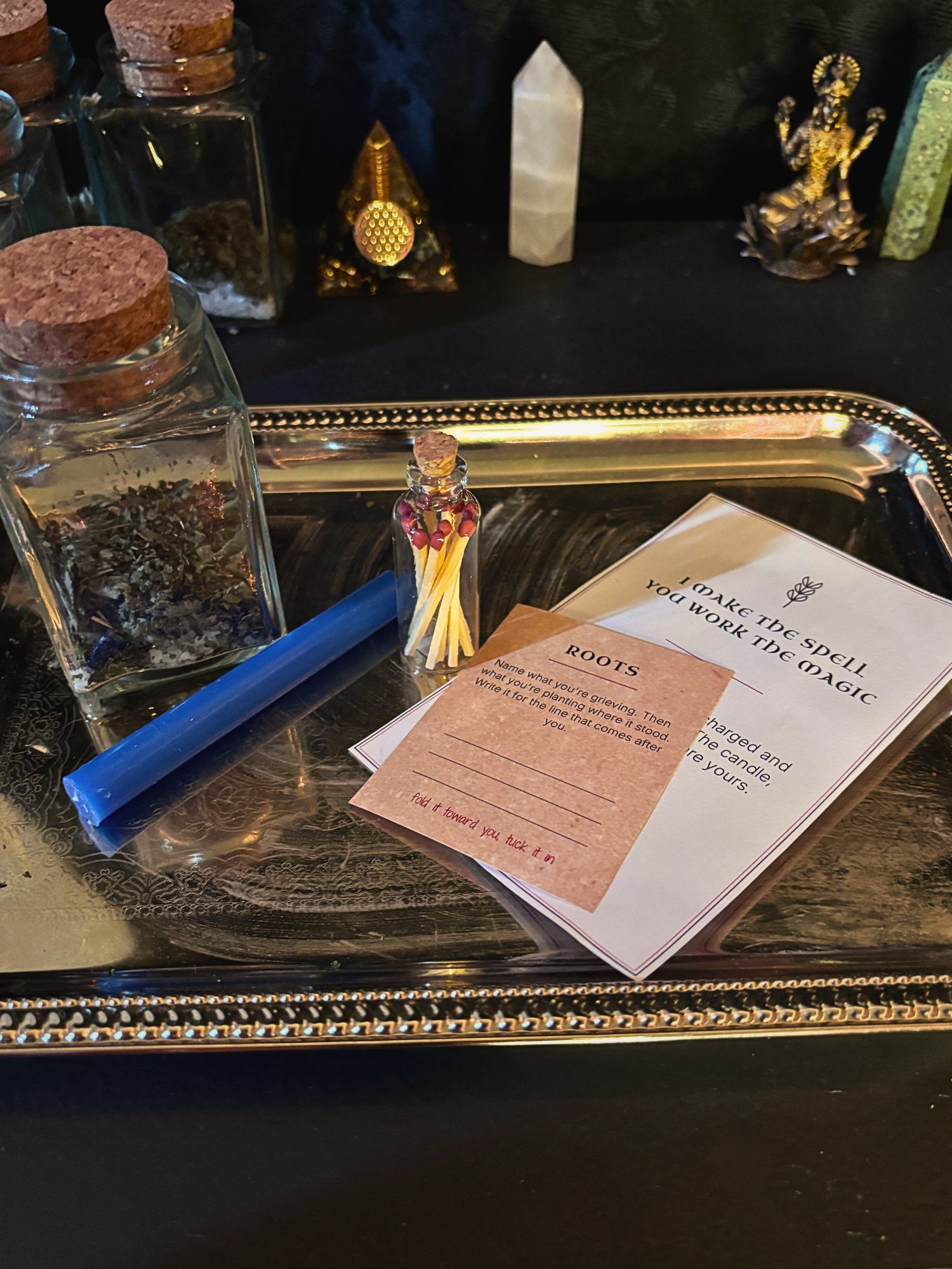 A ritual spell kit displayed on a silver tray, featuring a blue wand, a small glass jar of dried herbs, a matchstick holder with a miniature figure, and printed cards including one titled 'Irate the spell you work the Atoic'.
